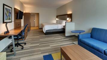 A hotel room with a bed, a desk with a chair, a sofa, a TV mounted on a wooden cabinet, and a small table.