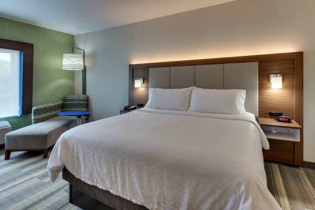 A hotel room with a large bed, a bedside table with a lamp, a chair, and a small table with a clock.