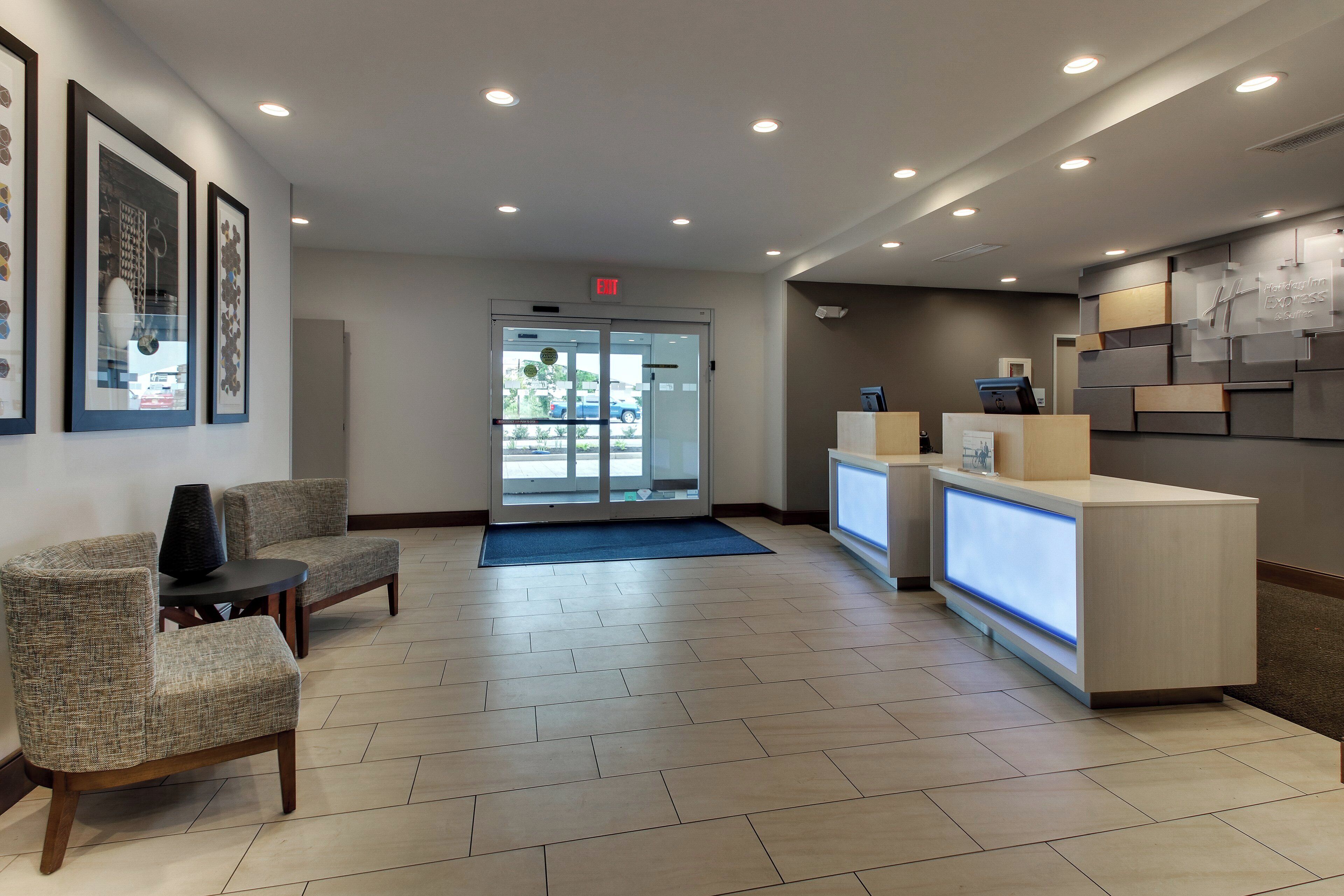 A hotel reception area with a reception desk, two chairs, a blue mat, and framed artwork on the wall.