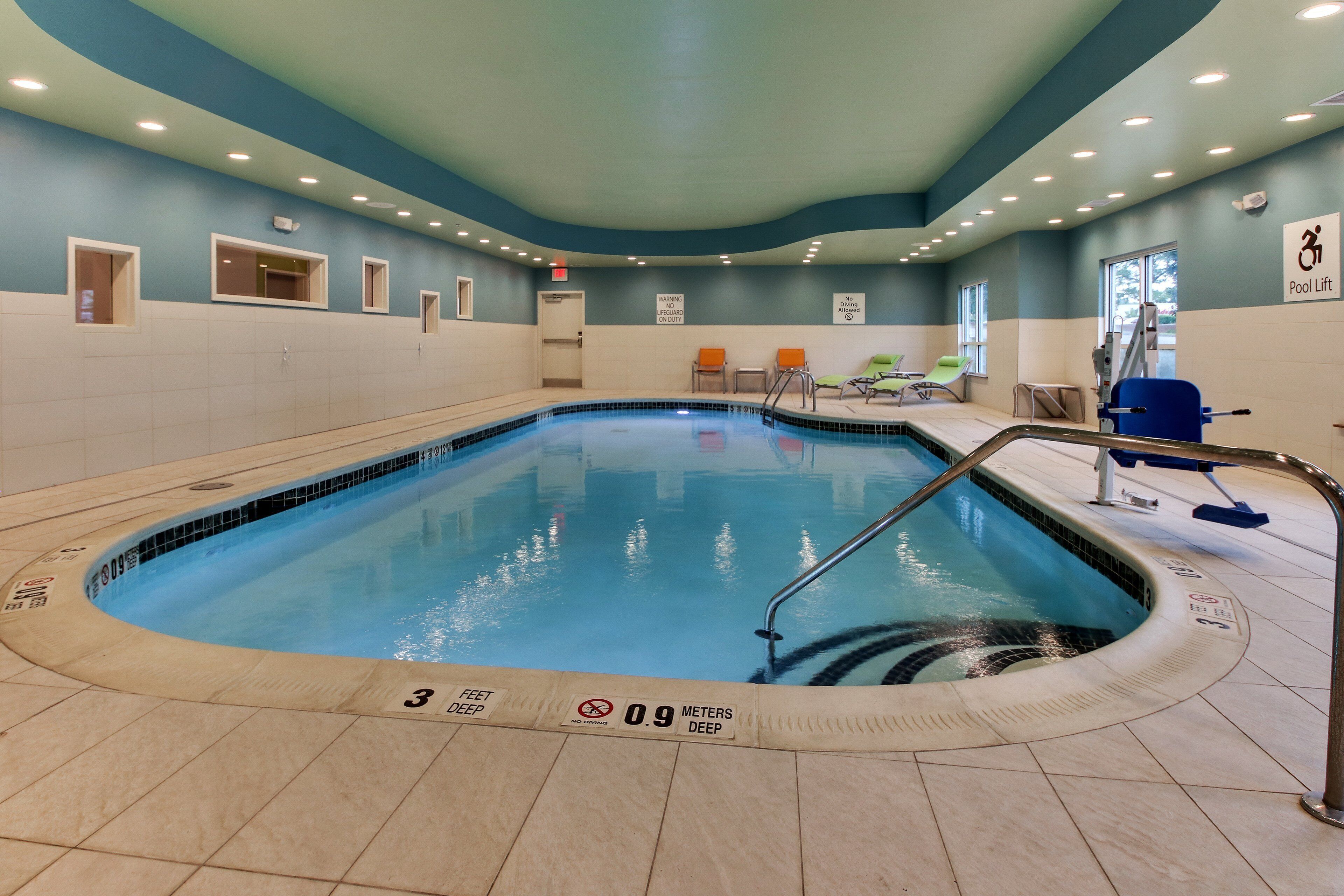 An indoor swimming pool with clear depth markers, a pool lift, and a seating area with chairs.