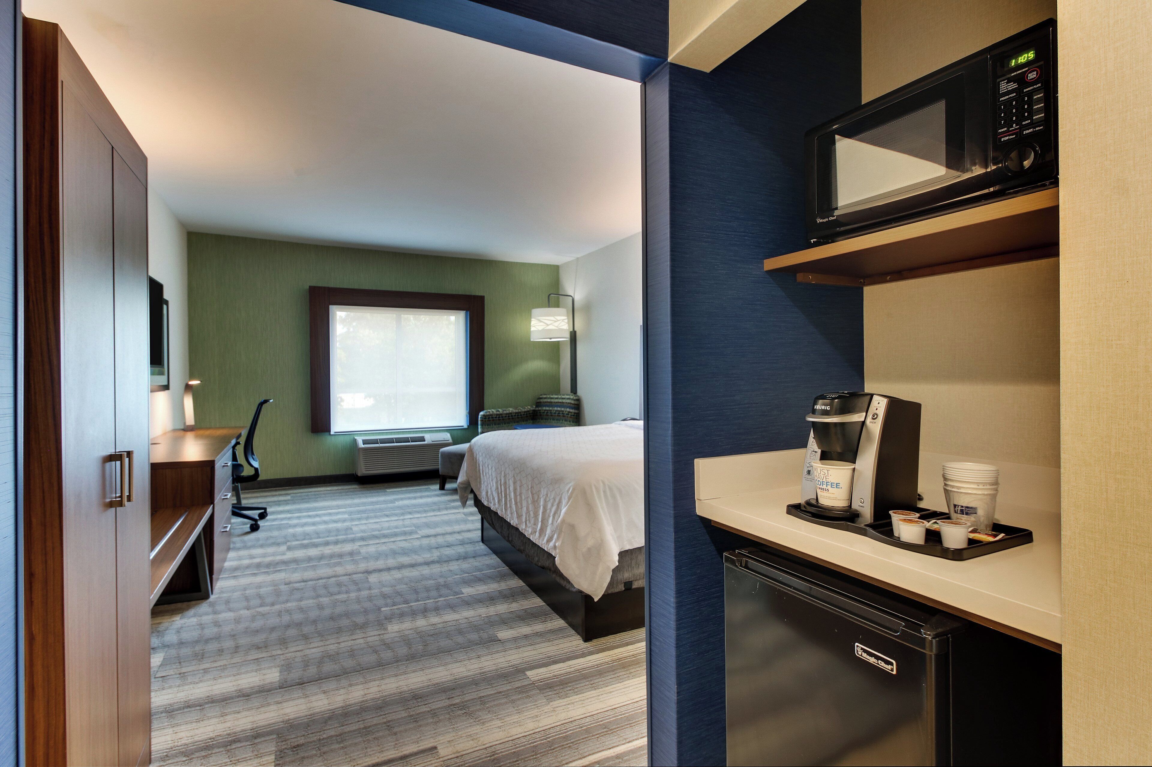 A hotel room with a bed, desk, microwave, coffee maker, and a view of a cityscape.