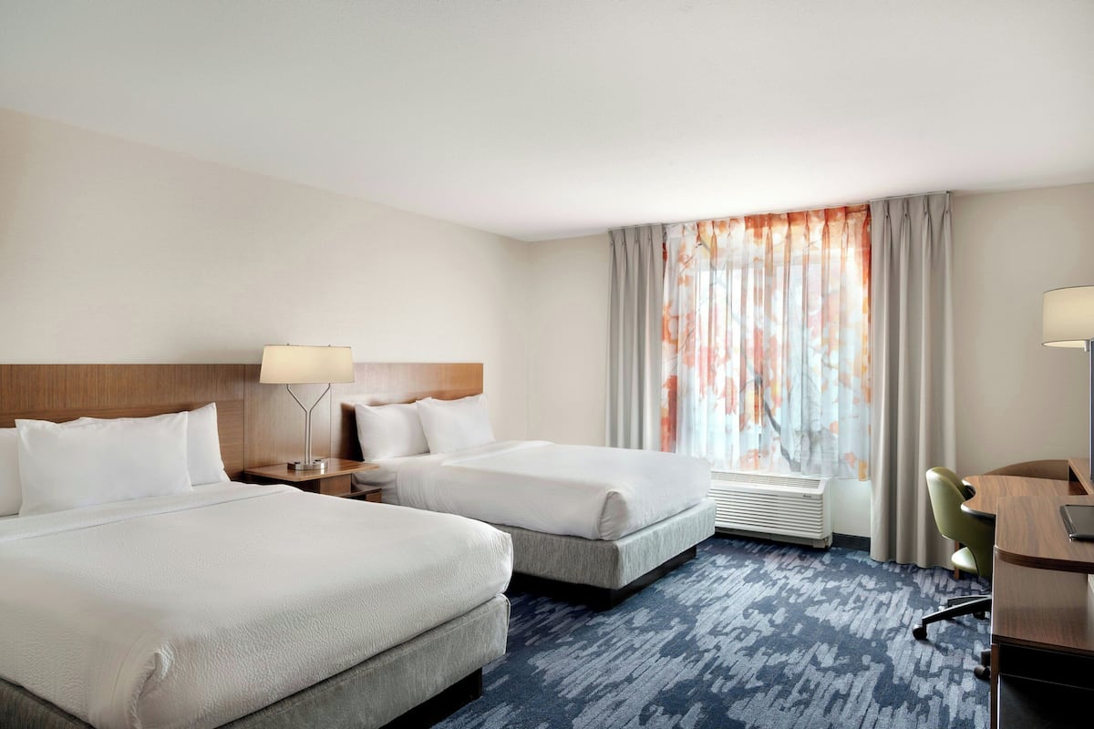 A hotel room with two beds, a desk, a chair, a lamp, and a window with curtains.