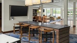 Un hall d’hôtel moderne doté d’un bar central, de différents espaces aménagés pour s’asseoir et d’un grand téléviseur à écran plat fixé au mur.