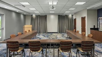 Un salón de conferencias con una larga mesa, sillas estampadas, un proyector, cortinas y vistas al exterior.