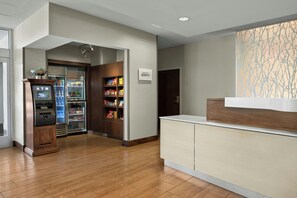 Eine Hotelhalle mit einem Geldautomaten, einem mit Snacks gefüllten Kühlschrank, einem Holzkabinett und einem Schild mit der Wegbeschreibung.