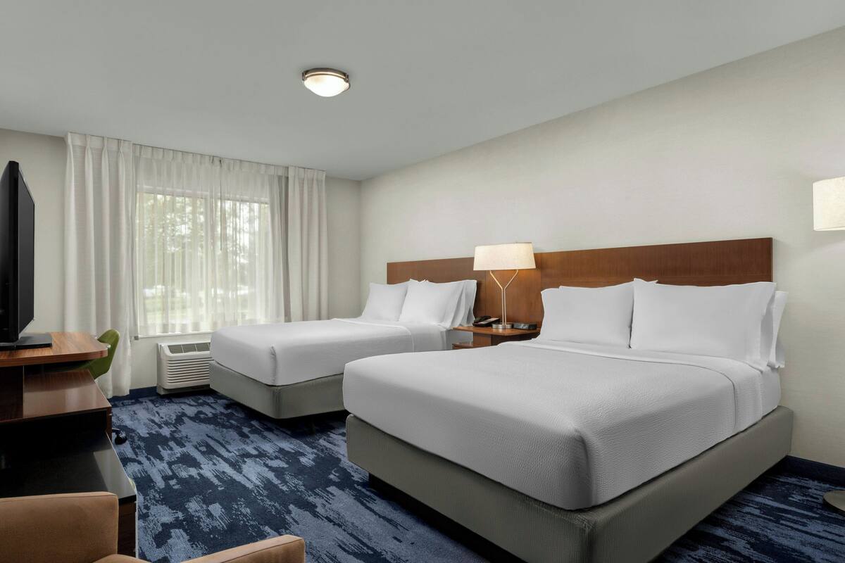 A hotel room with two beds, a desk with a TV, a lamp, and a large window with curtains.