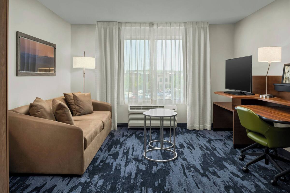 A hotel room with a sofa, a small round table, a desk with a computer and TV, and a window with curtains.