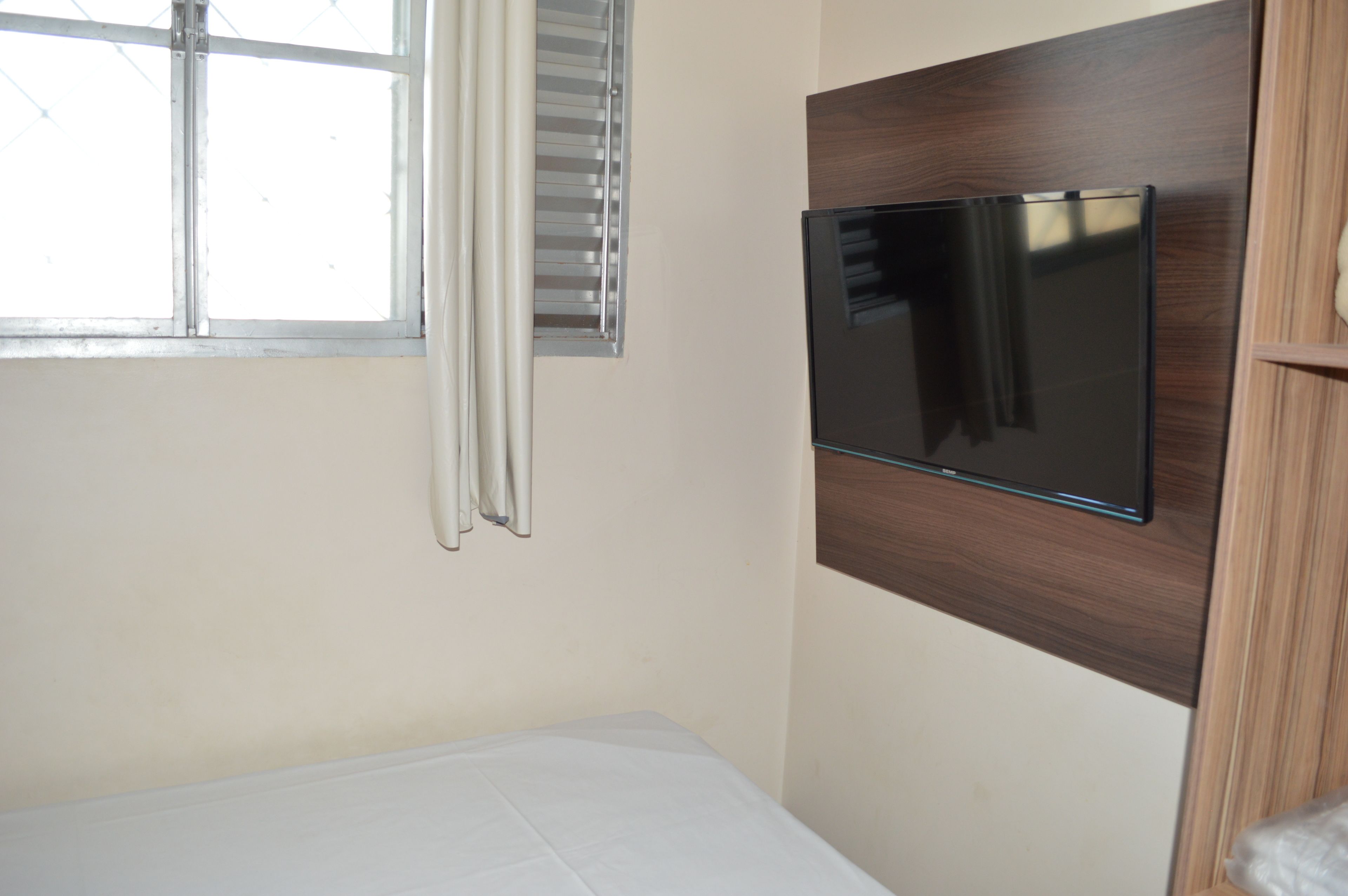 Quarto com cama, TV fixada na parede, janela com cortinas e painel de madeira na parede.