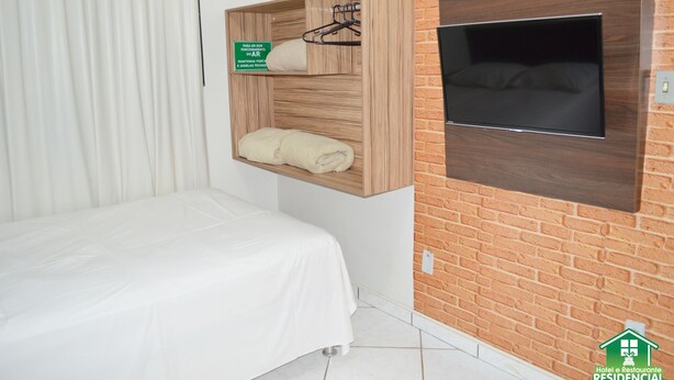 A hotel room with a bed, a wooden shelf with a towel and a hanging item, and a TV mounted on a brick wall. - Hotel e Restaurante Residencial (Montes Claros)
