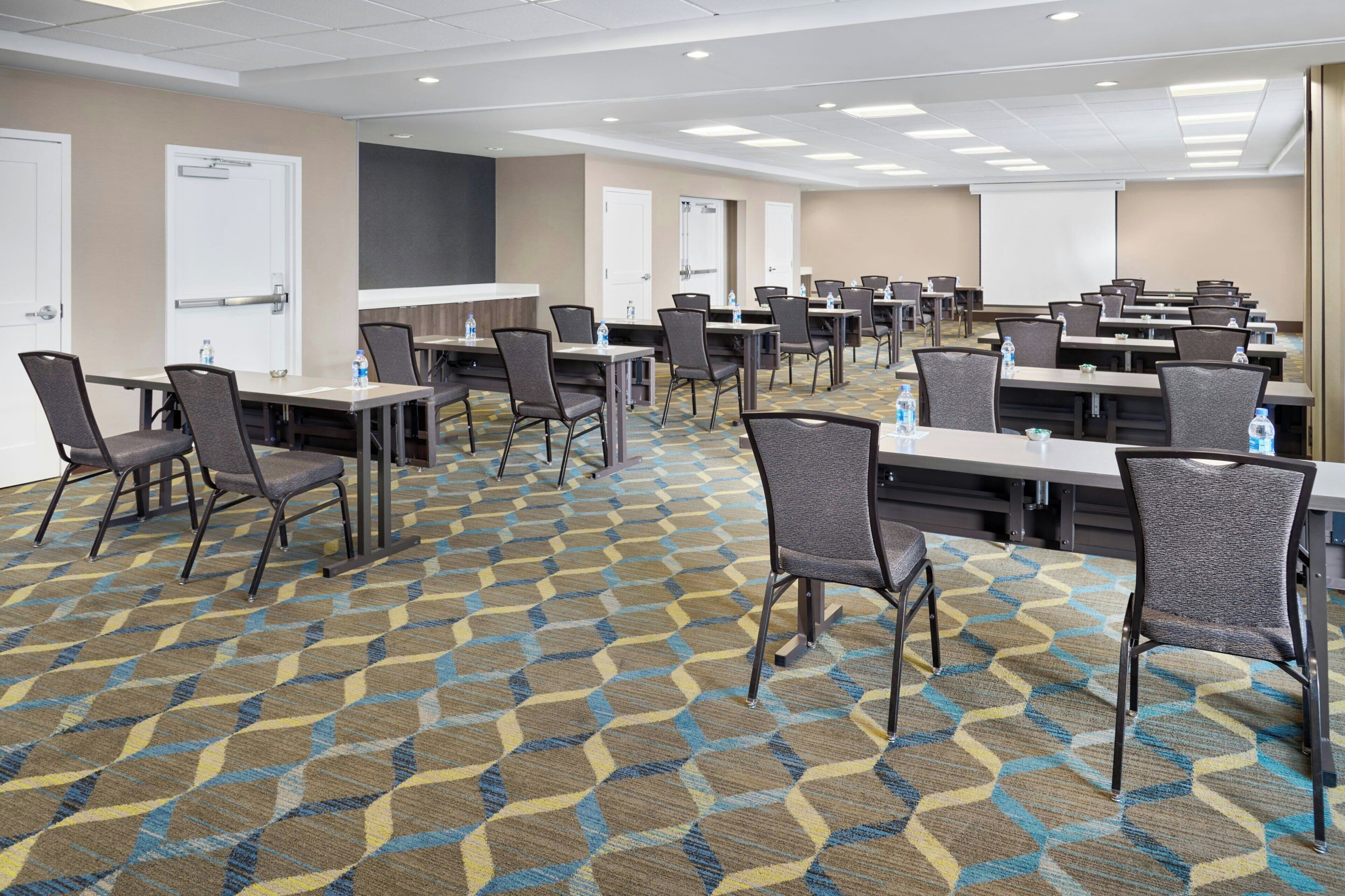A conference room with multiple tables and chairs, a projector screen, and a carpeted floor.