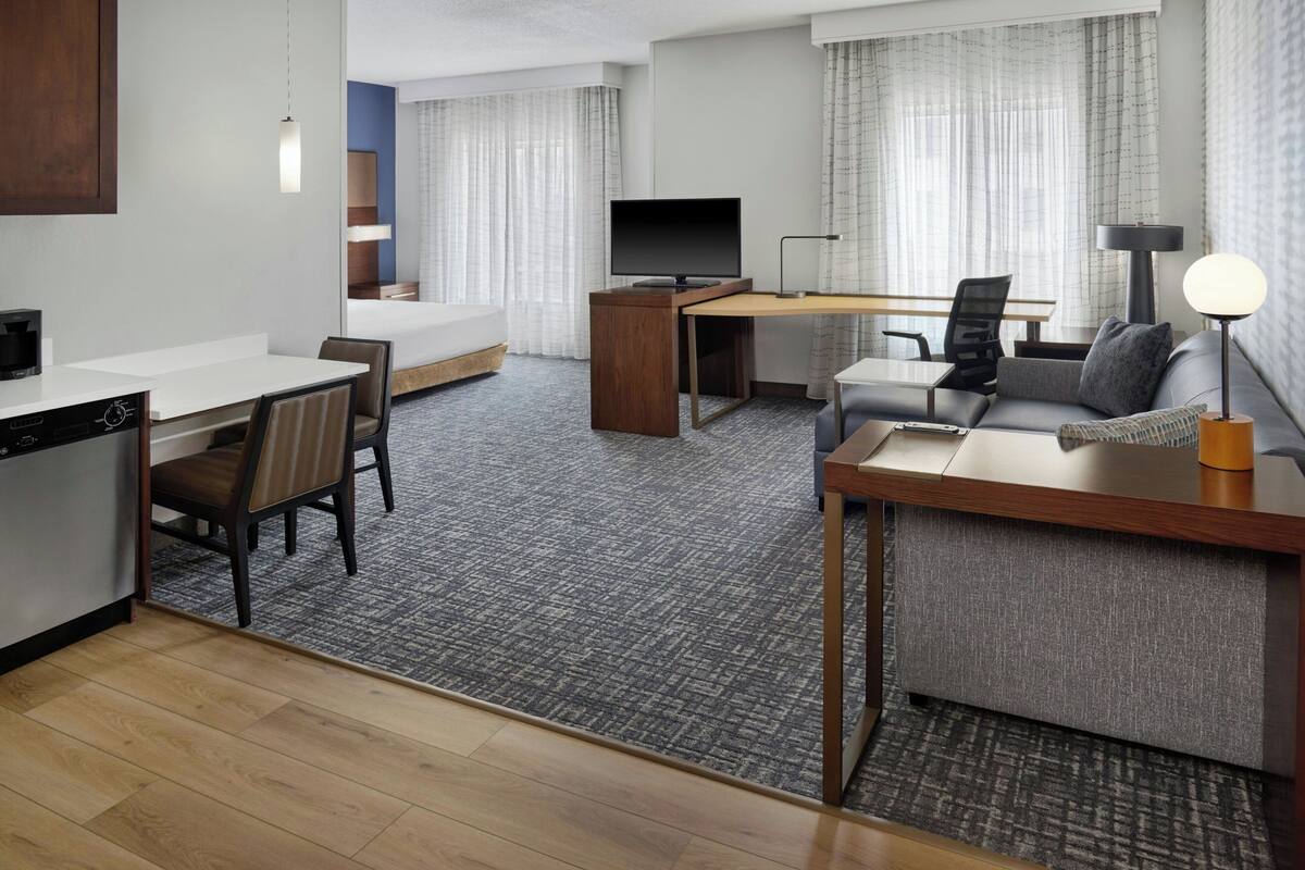 A modern hotel room with a kitchenette, a sofa, a desk, and a TV.