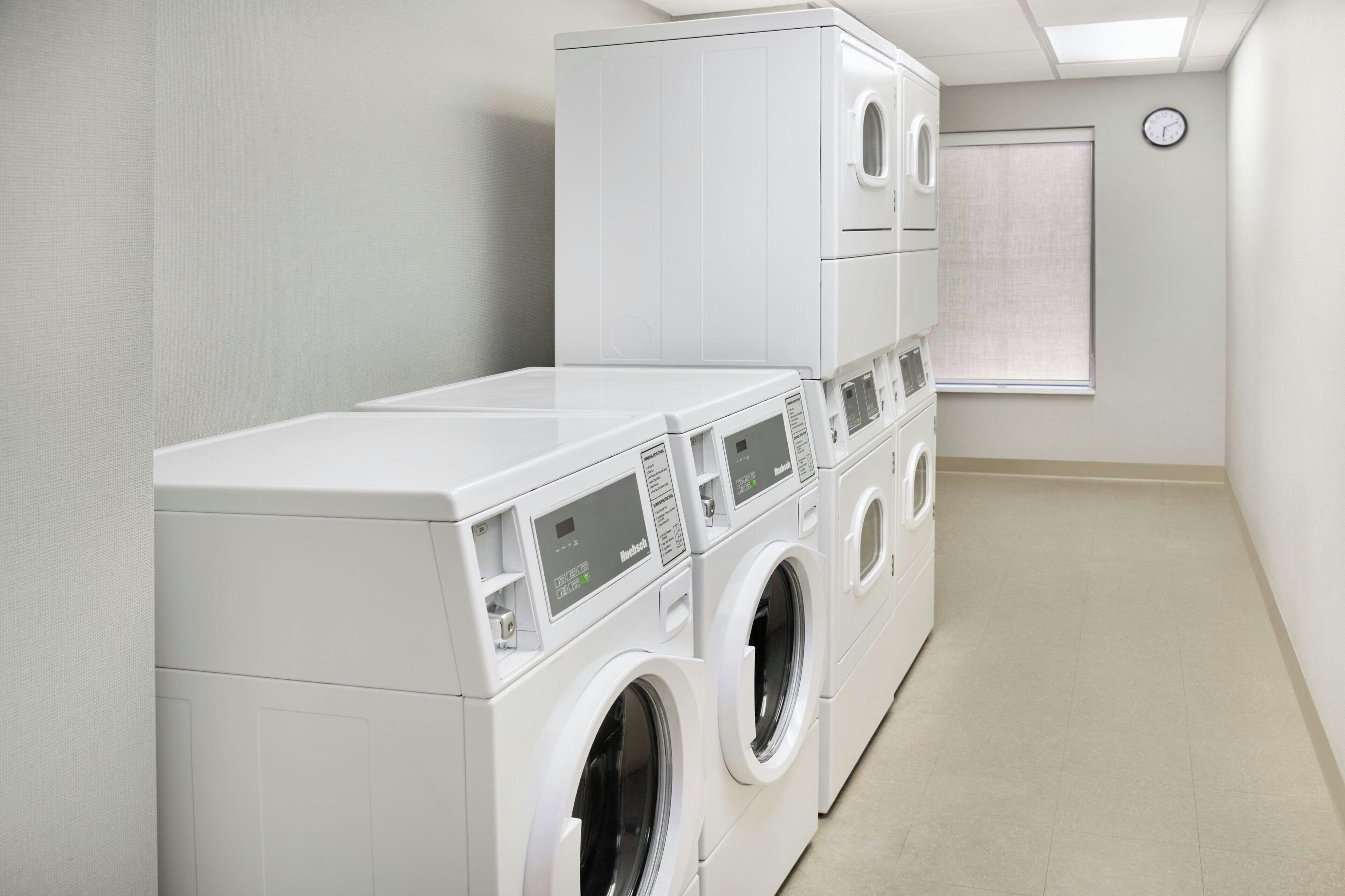A laundry room with stacked washers and dryers, a clock on the wall, and a window with blinds.