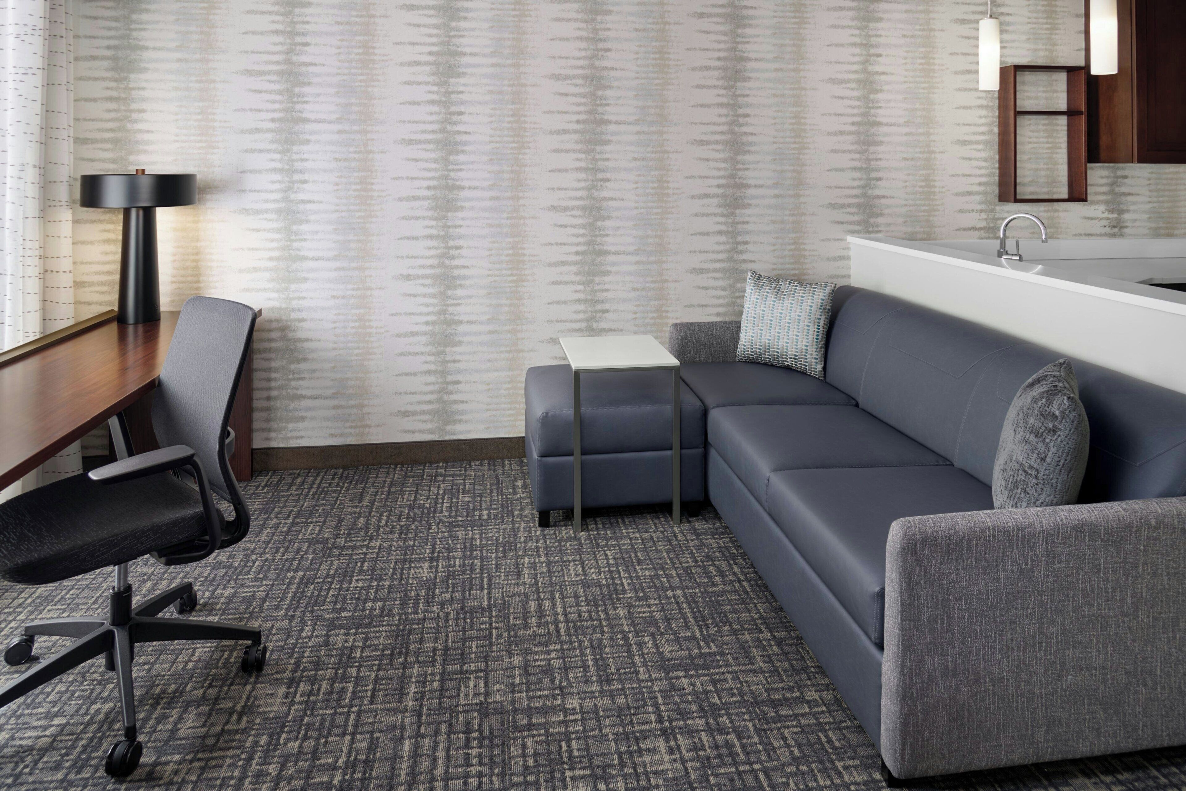 A modern hotel room with a gray carpet, a gray sofa, a small side table, a desk with a chair, and a large window.