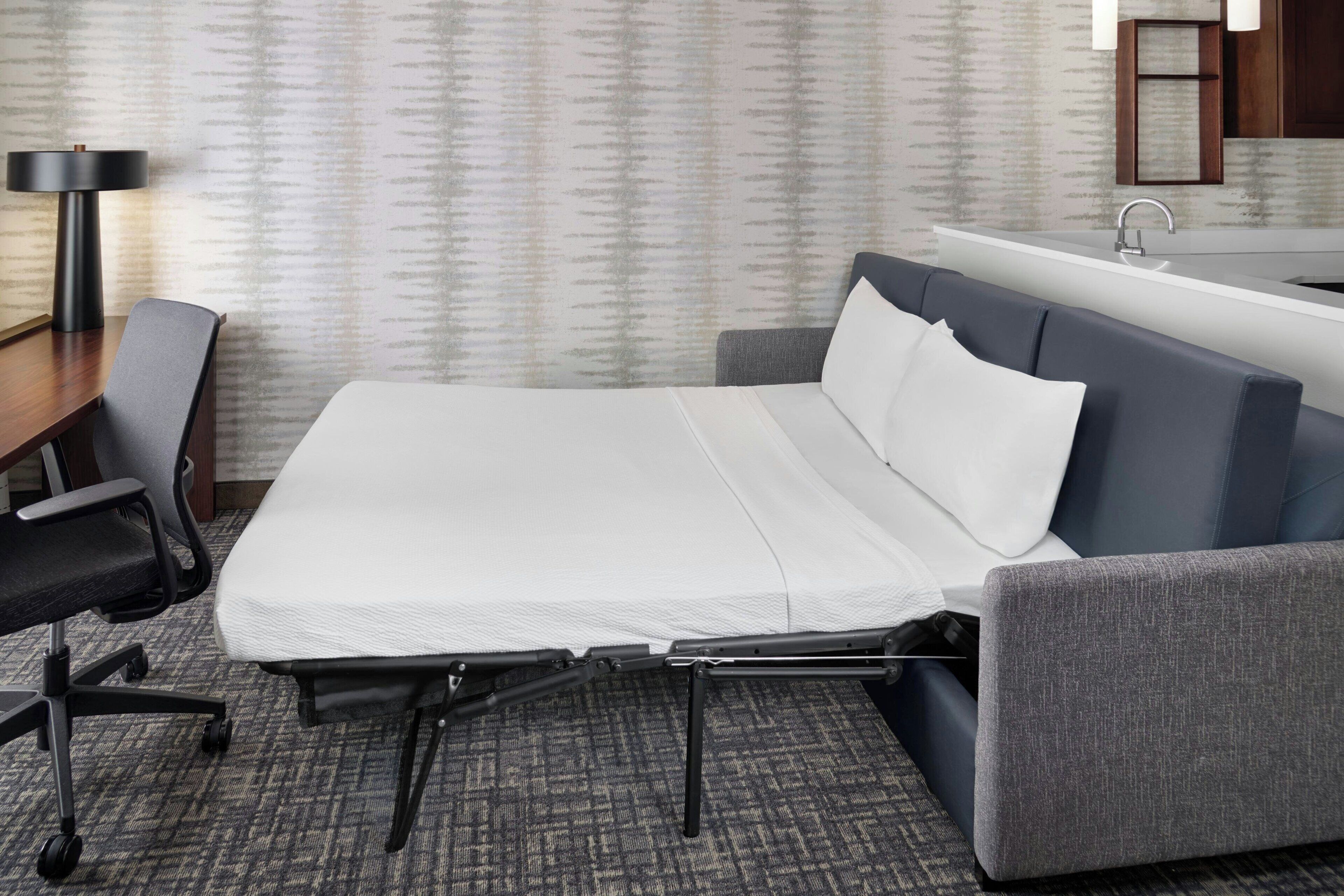 Une chambre d’hôtel de taille réduite, comprenant un lit, un bureau, une chaise, une lampe et une salle de bain avec un lavabo.