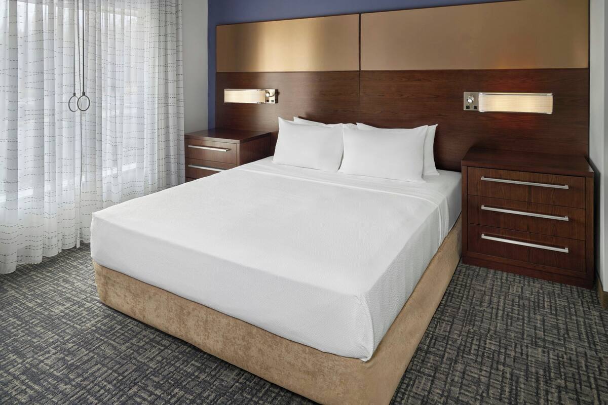 A hotel room with a large bed, bedside tables, a nightstand, and a headboard with integrated lighting.