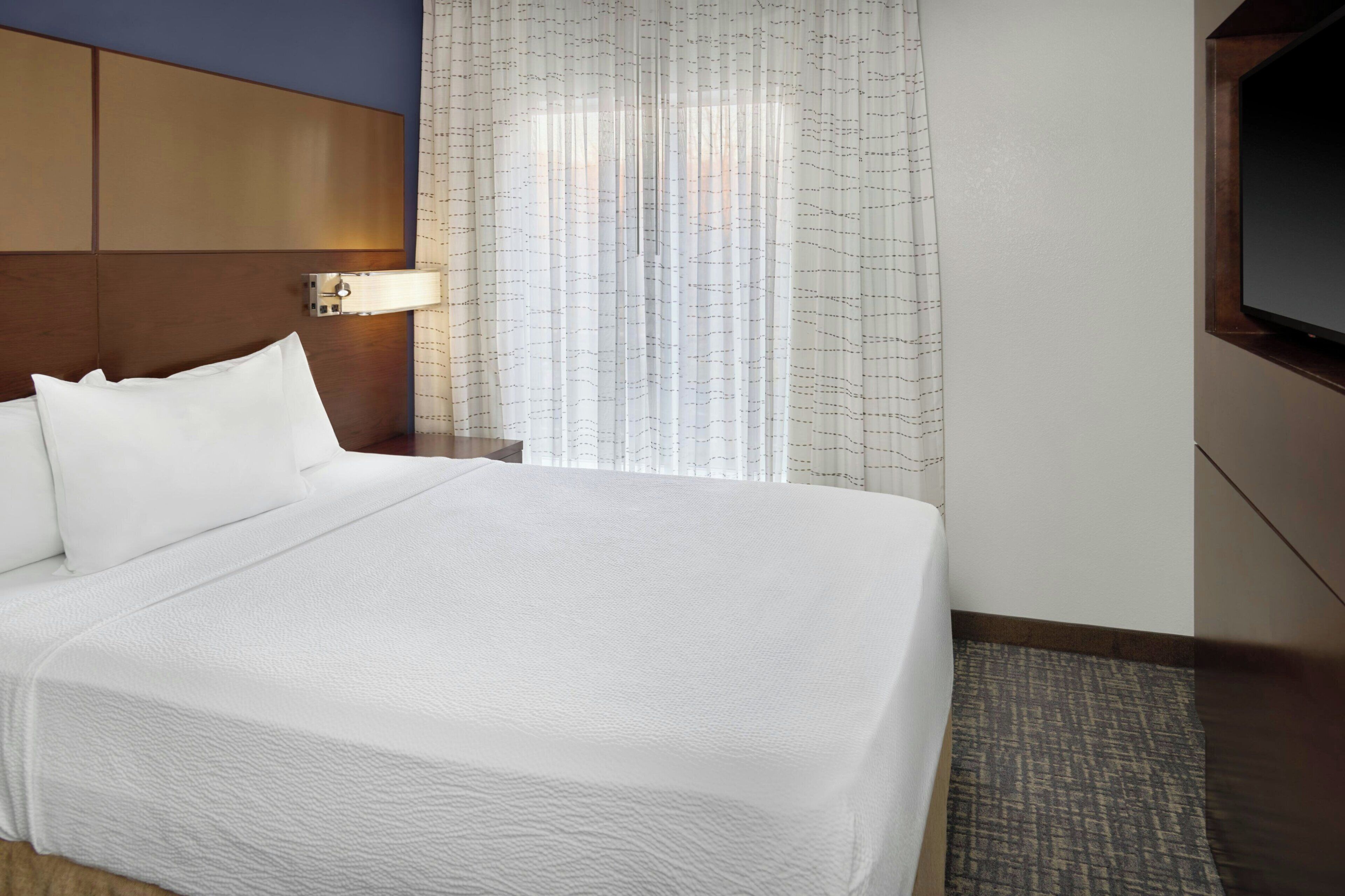 A hotel room with a large bed, a nightstand with a lamp, a TV mounted on the wall, and a window with curtains.