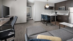 Ein modernes Hotelzimmer mit einer Couch, einem Essbereich mit Stühlen, einem Fernseher und einer Küchenzeile inklusive Mikrowelle und Kühlschrank.
