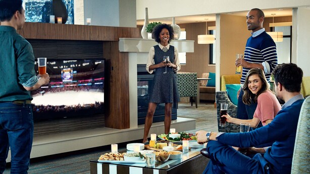 Eine moderne Hotelhalle mit einem großen Flachbildfernseher, einem Buffet mit Speisen und Getränken sowie einer Sitzecke mit Sofas und einem Couchtisch.
