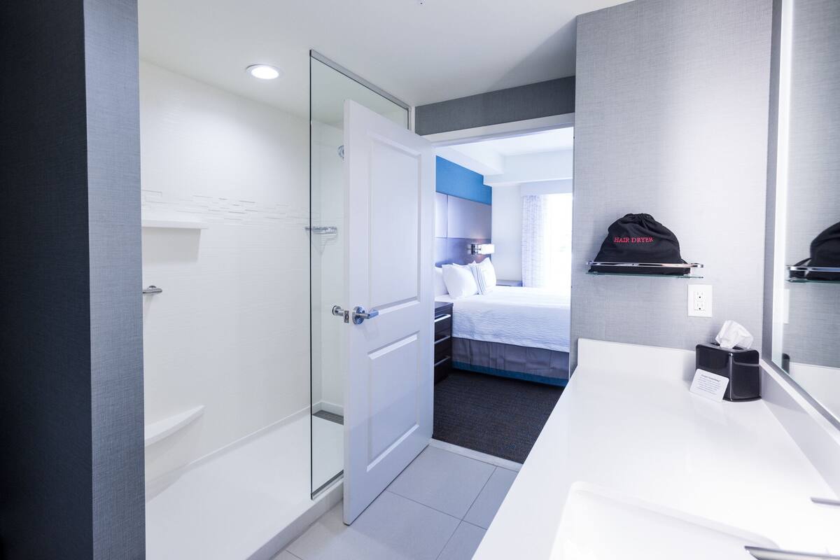 Camera d'albergo con porta doccia in vetro, un letto con testiera blu, un mobiletto del bagno bianco e un cappello nero appeso al muro.