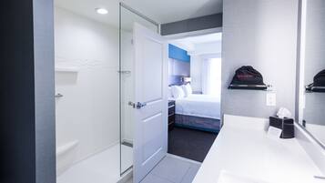 Una habitación de hotel con mampara de vidrio en la ducha, una cama con cabecera azul, un mueble de baño blanco y un sombrero negro en la pared.
