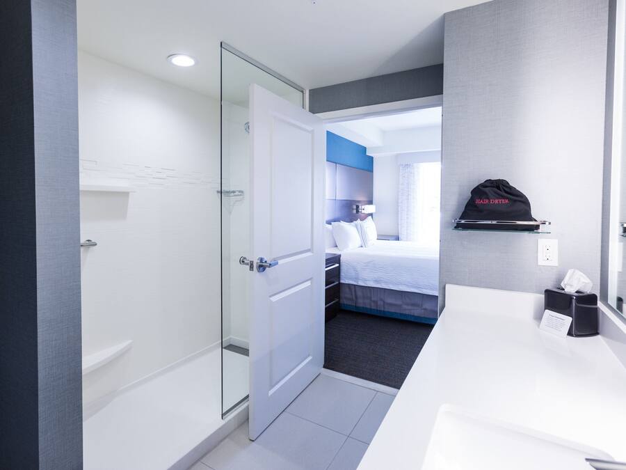 Una habitación de hotel con mampara de vidrio en la ducha, una cama con cabecera azul, un mueble de baño blanco y un sombrero negro en la pared.