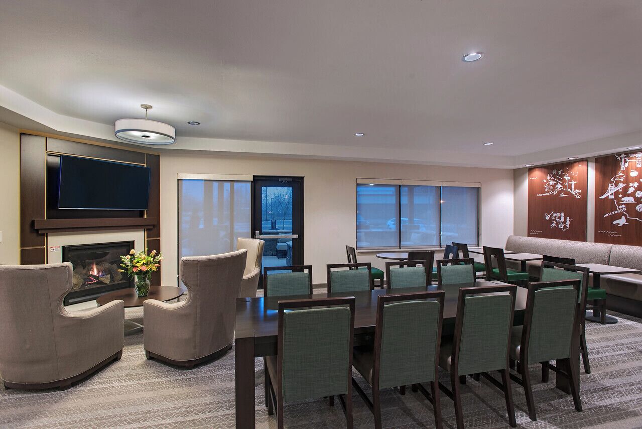 A modern hotel room with a fireplace, a flat-screen TV, a dining area with chairs and tables, and large windows offering a view of the city.