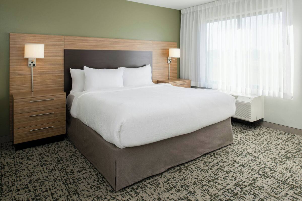 A hotel room with a large bed, two bedside tables with lamps, a window with sheer curtains, and a patterned carpet.