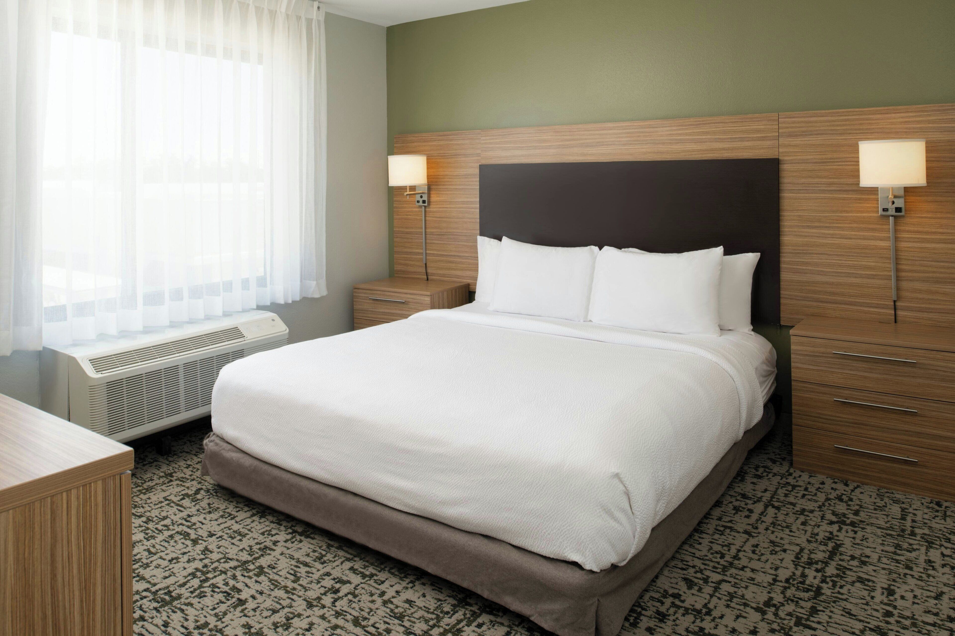 Quarto de hotel com uma cama grande, duas mesas de cabeceira, uma mesinha de apoio, uma escrivaninha e uma janela com cortinas leves.
