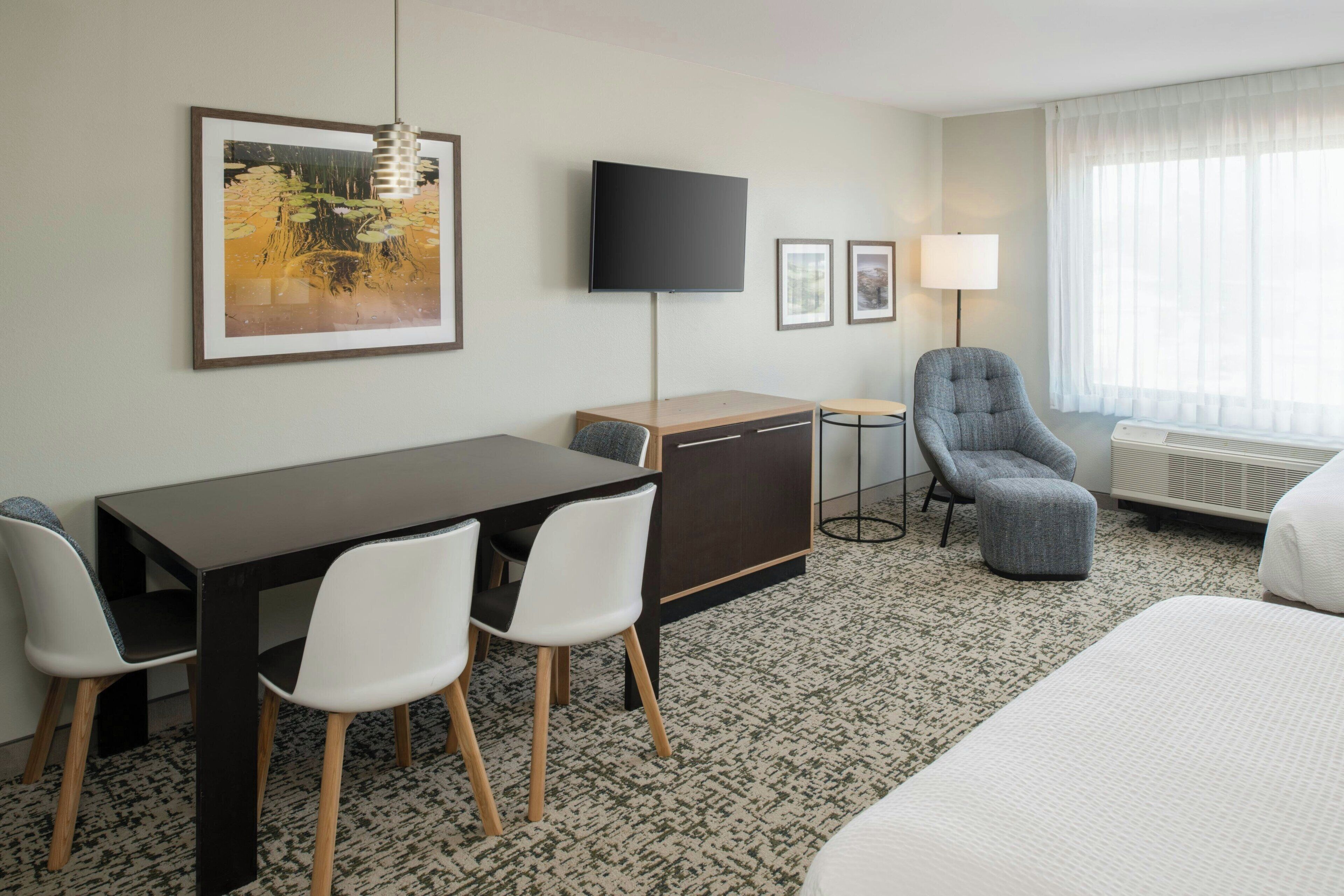 Habitación de hotel con cama, escritorio con sillas, televisión, lámpara y un amplio ventanal con cortinas.