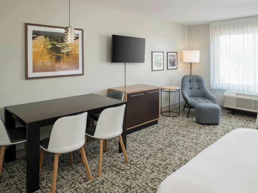 Habitación de hotel con cama, escritorio con sillas, televisión, lámpara y un amplio ventanal con cortinas.