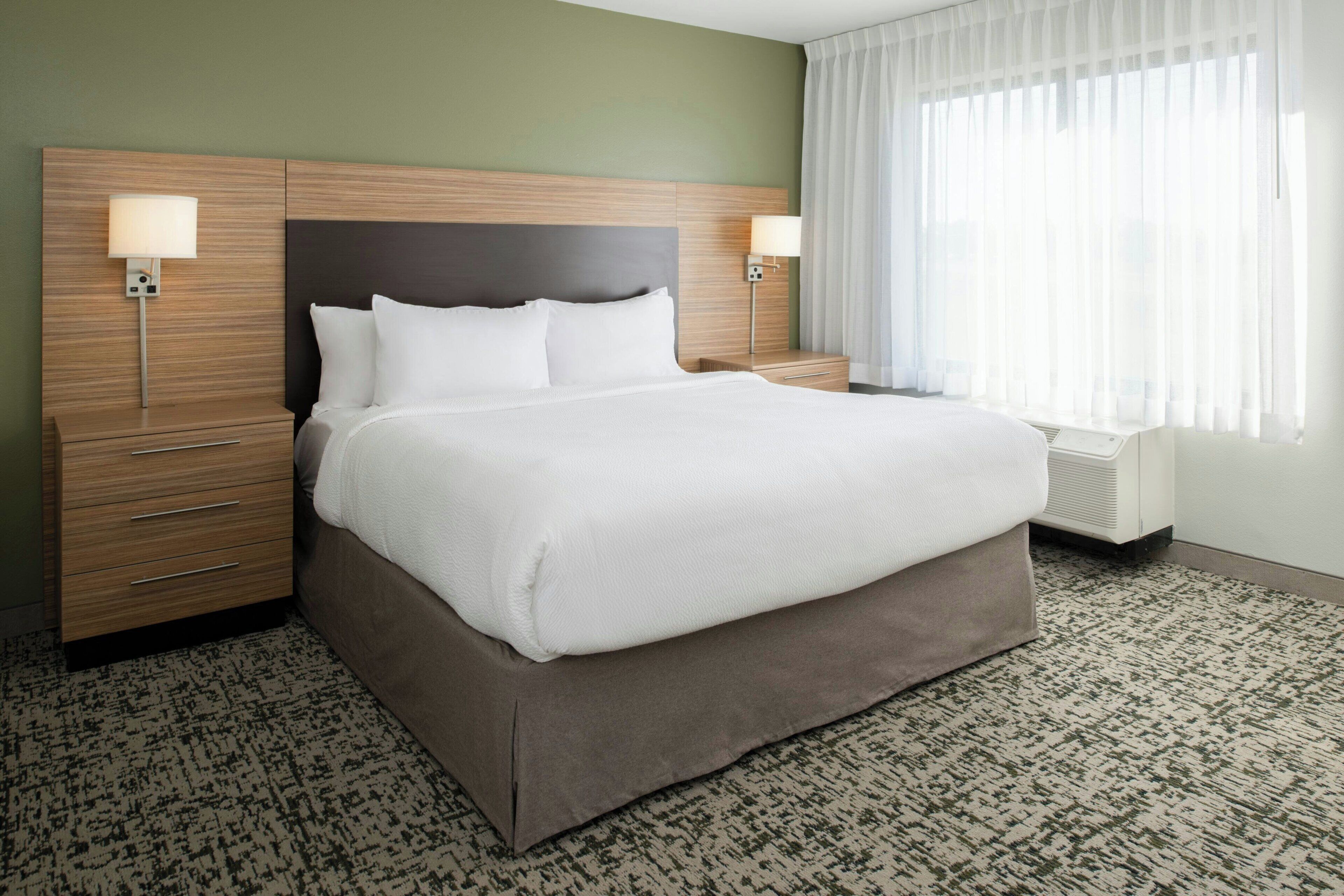 A hotel room with a large bed, two bedside tables, a nightstand, and a window with sheer curtains.