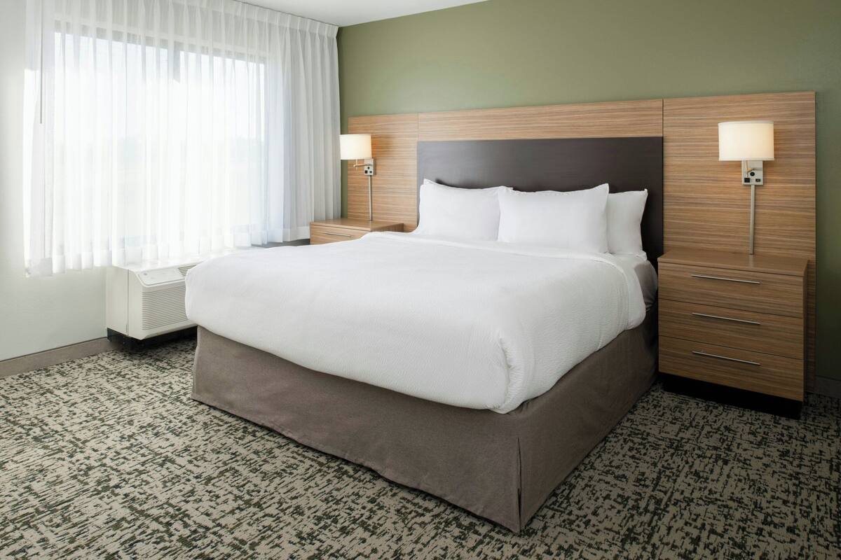 A hotel room with a large bed, wooden headboard, two bedside tables with lamps, a window with sheer curtains, and a patterned carpet.