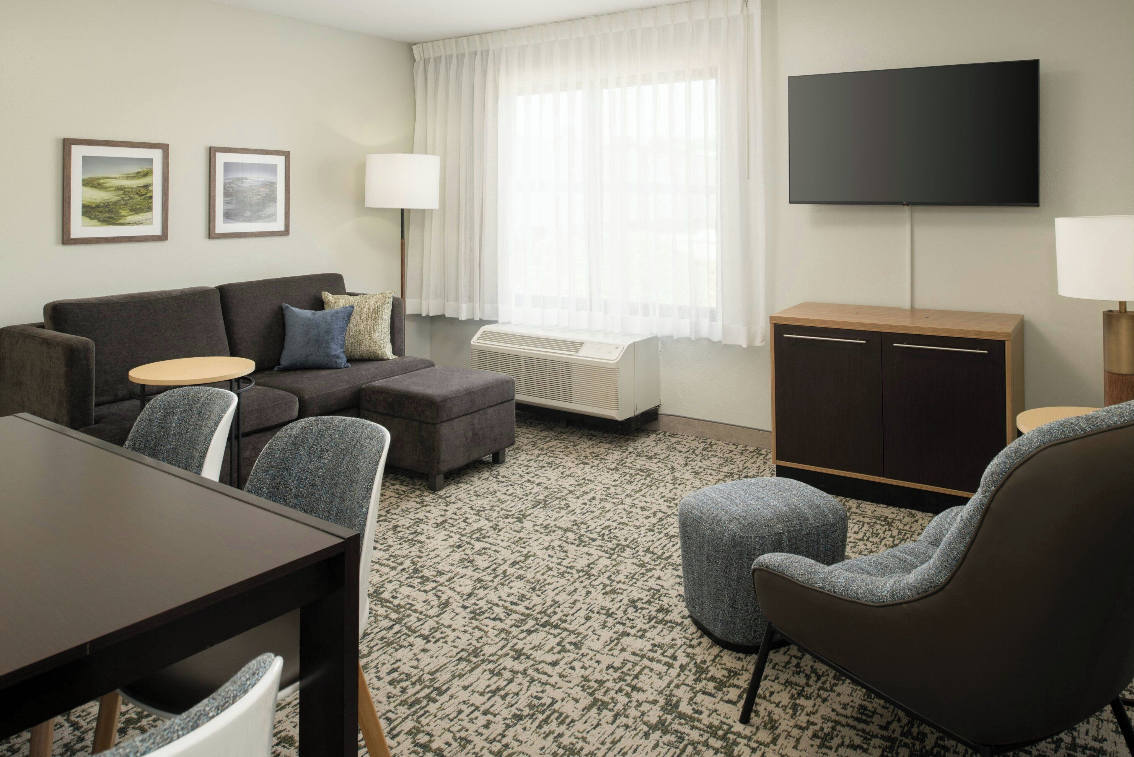 A modern hotel room with a sofa, armchairs, a dining table, a flat-screen TV, and a cabinet.