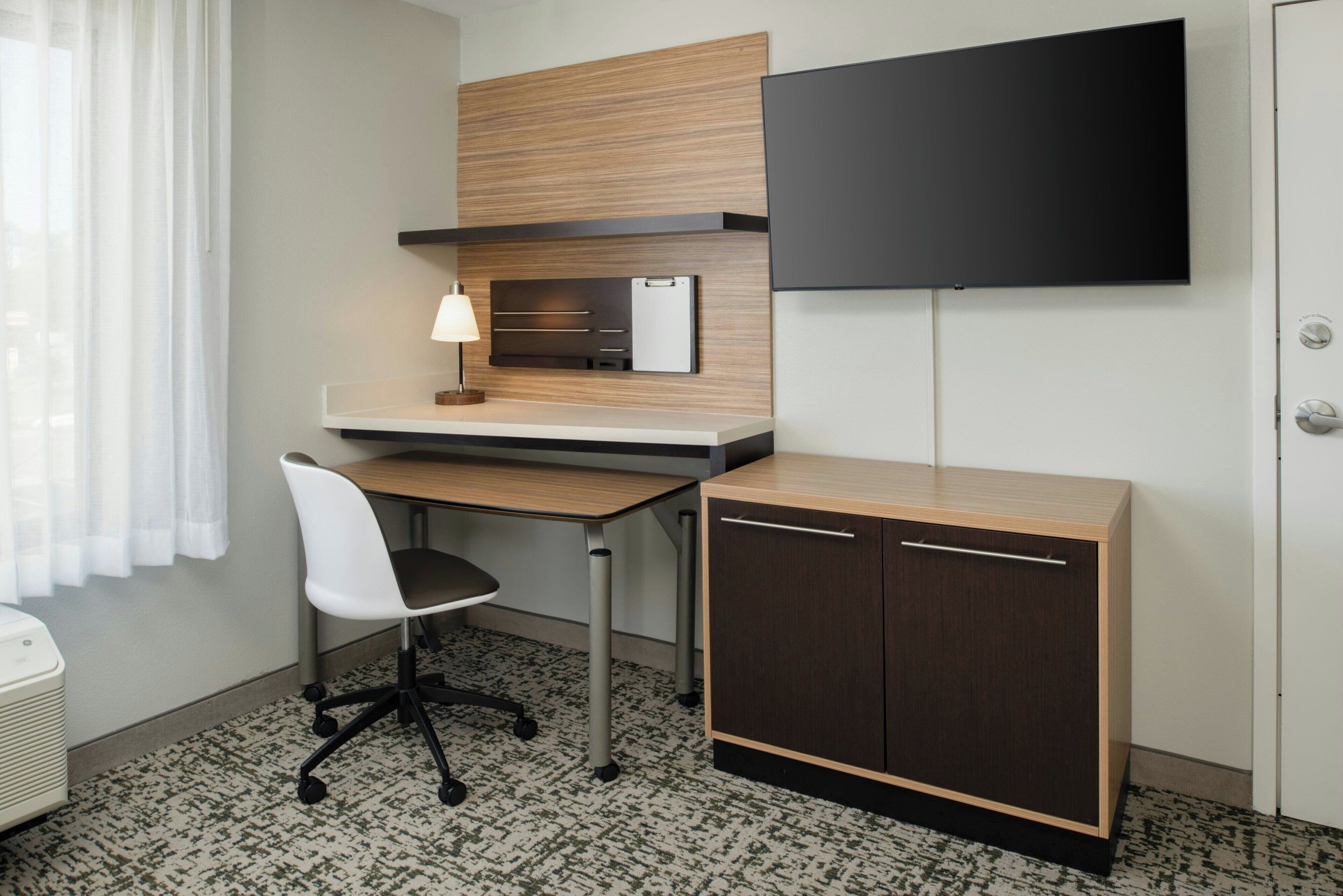 A modern hotel room with a wooden desk, a chair, a flat-screen TV, a cabinet, and a window with curtains.