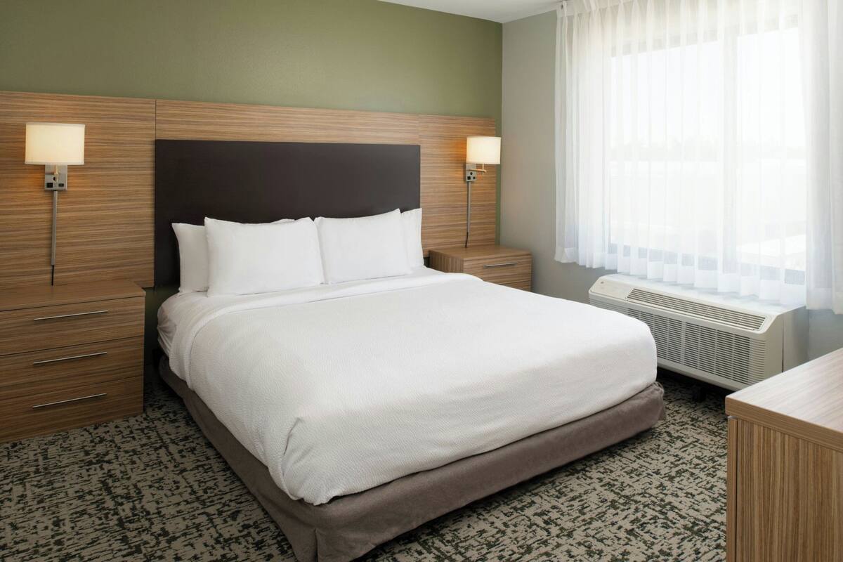 A hotel room with a large bed, two bedside tables, a nightstand, a desk, and a window with sheer curtains.