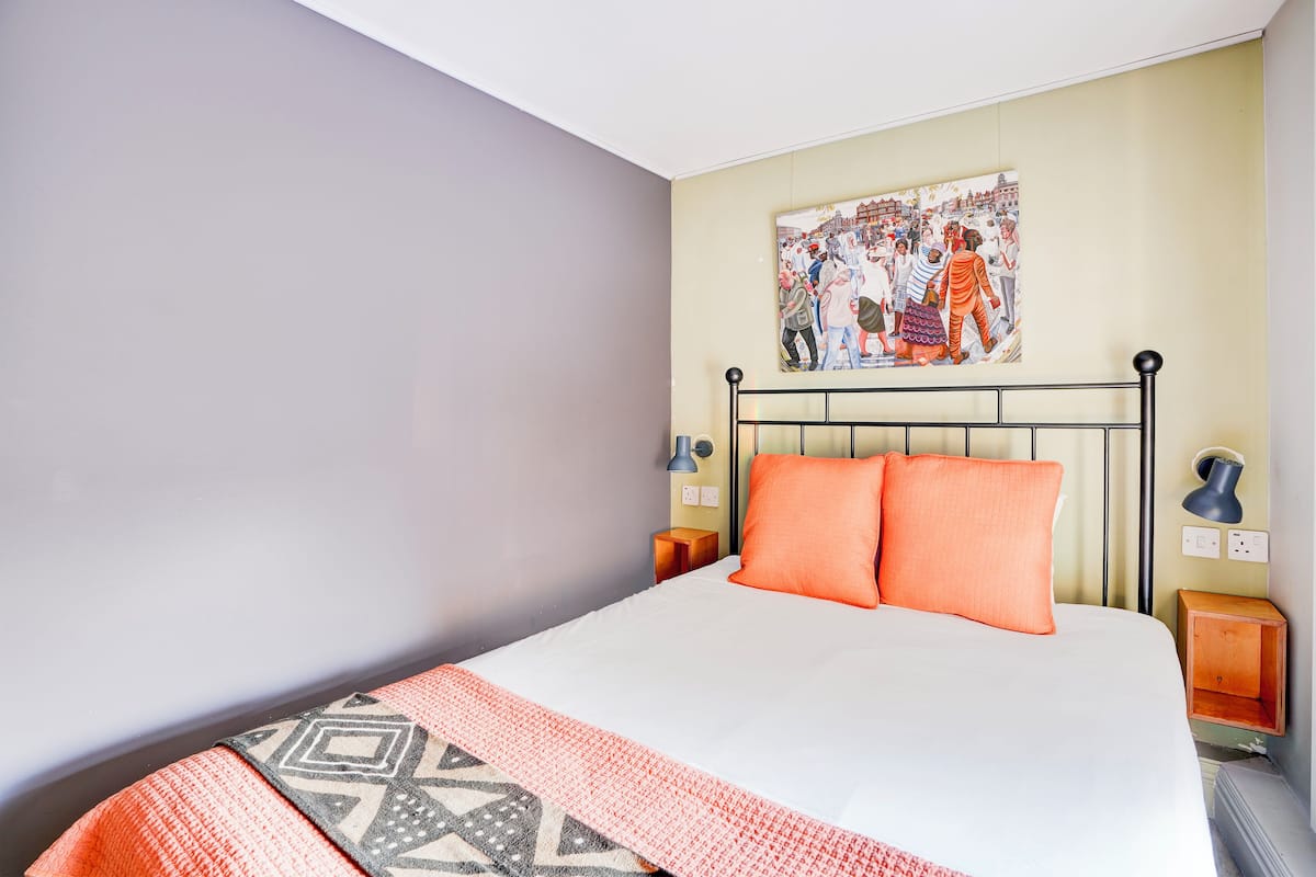 A neatly made bed with two orange pillows, a patterned throw, and a wall-mounted painting depicting a group of people.