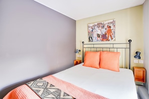 Quarto com cama, duas almofadas, mesa de cabeceira, abajur e pintura pendurada na parede.