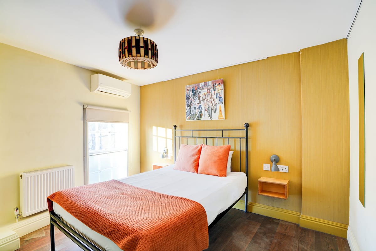A bedroom with a bed, an air conditioner, a radiator, a window, and a wall-mounted shelf.