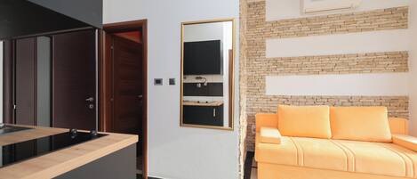 Una cocina moderna con estufa integrada, una puerta de madera y un lavamanos pequeño. Hay un sofá con cojines naranjas frente a una pared con diseño de ladrillo.
