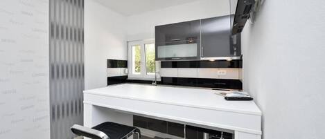 Een moderne keuken met een wit aanrecht, zwarte kasten en roestvrijstalen apparatuur. Er staan twee barkrukken met zwarte zittingen en chromen poten.