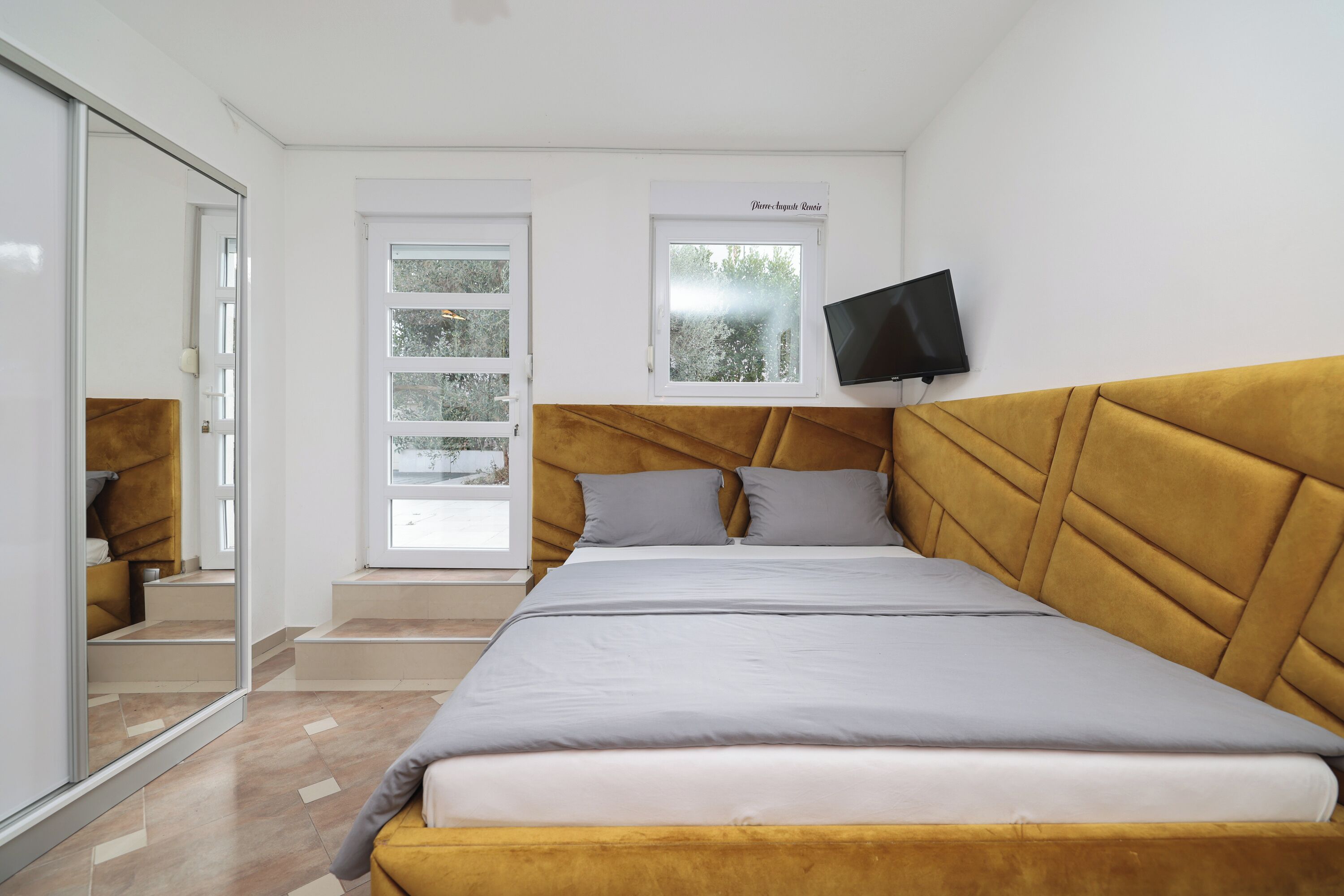 A bedroom with a large bed, a TV mounted on the wall, and a window with a view of trees.