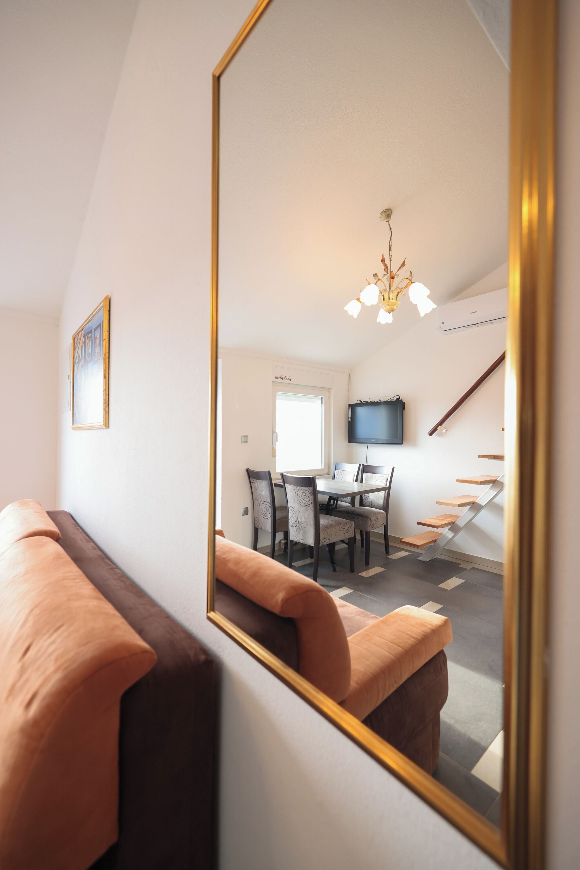 A modern living room with a large mirror reflecting a dining area, a staircase, and a television mounted on the wall.