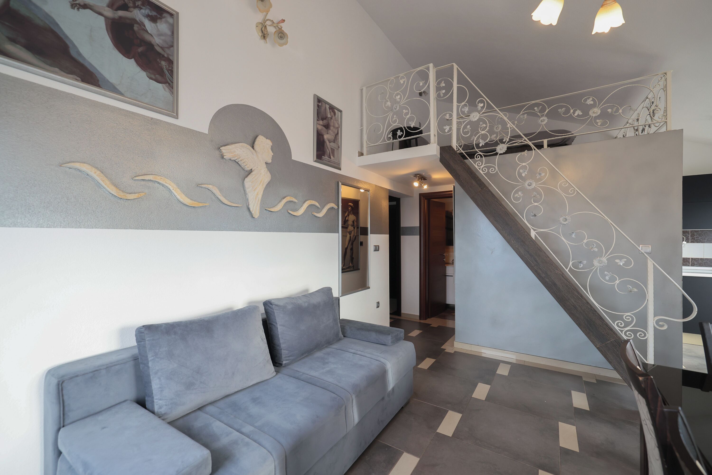 A modern living room with a grey sofa, a staircase with decorative railings, and artwork on the walls.