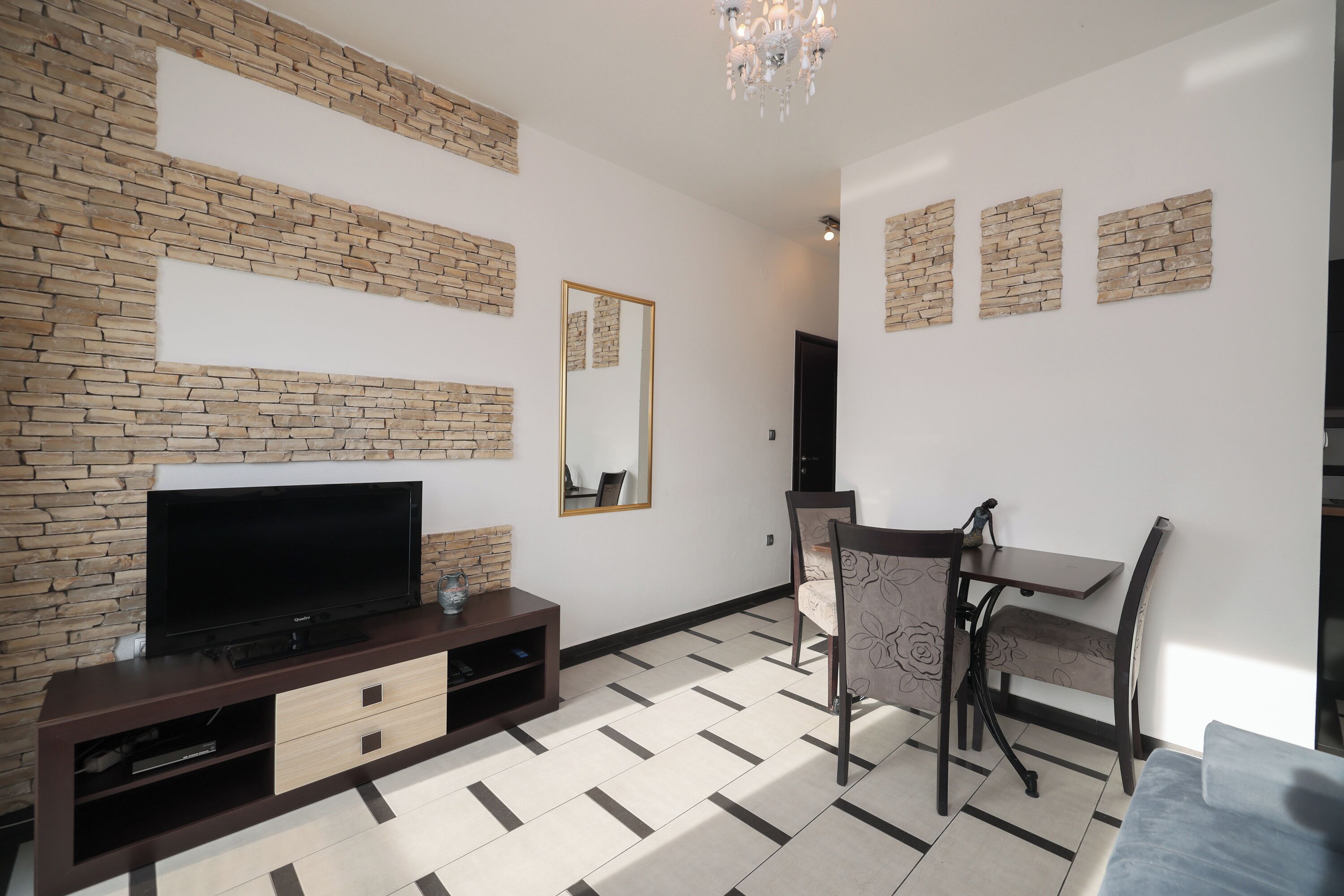 A modern living room with a flat-screen TV on a dark wooden entertainment unit, a dining area with a table and chairs, and a patterned rug on the floor.
