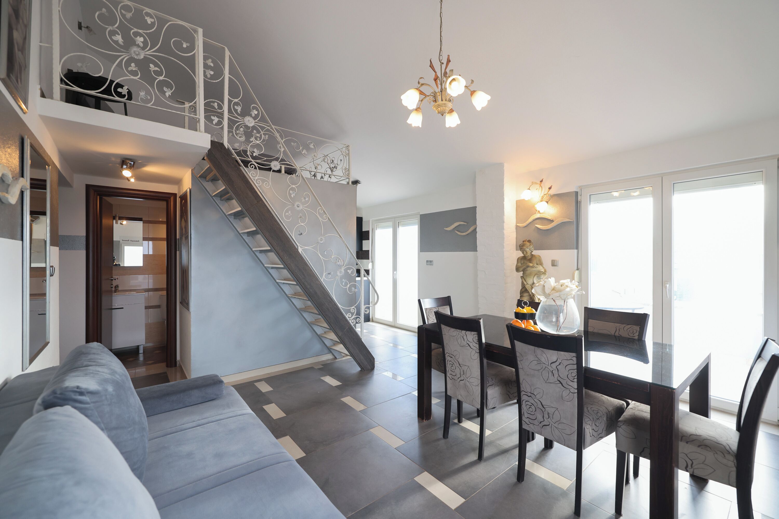 Un salon moderne avec un canapé gris, une table à manger avec des chaises, un escalier doté de rampes décoratives et un lustre.