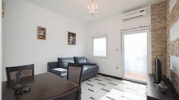 Una sala moderna con una mesa de comedor, un sofá y una araña. Hay una puerta que da a un balcón y un televisor sobre un mueble.