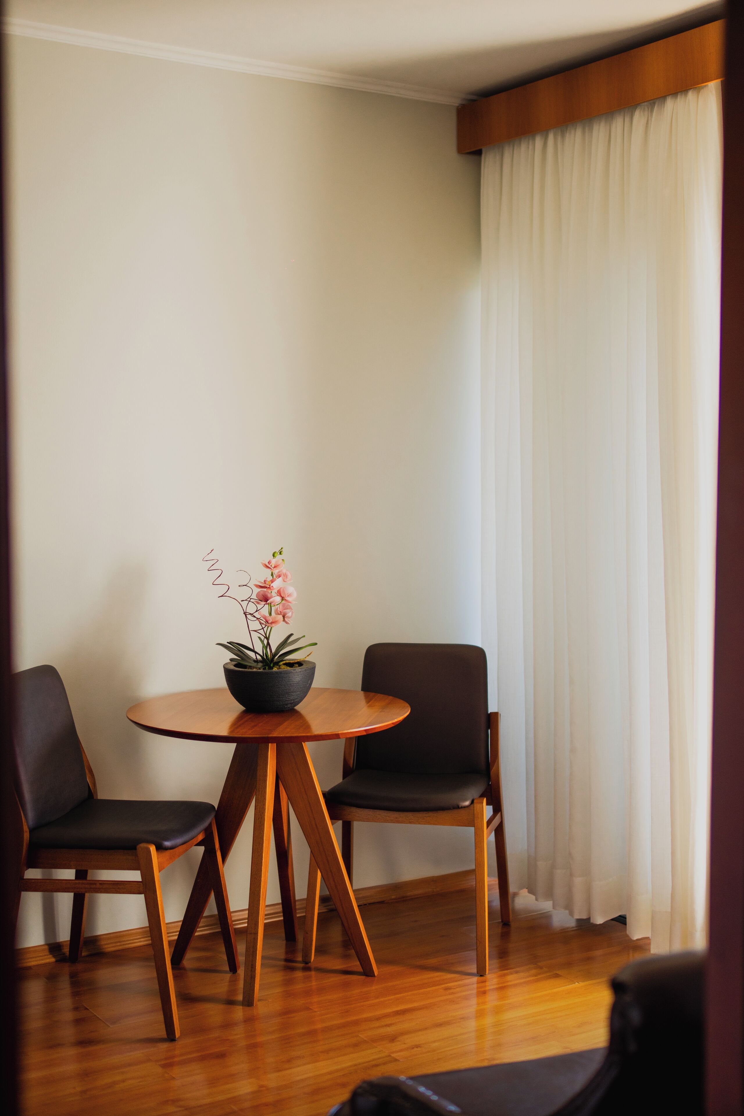 A room with a round wooden table, two chairs, a potted plant, and a white curtain.