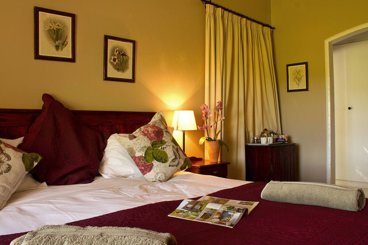 A neatly made bed with a red and white floral bedspread, a magazine on the bed, and a nightstand with a lamp and a vase of flowers.