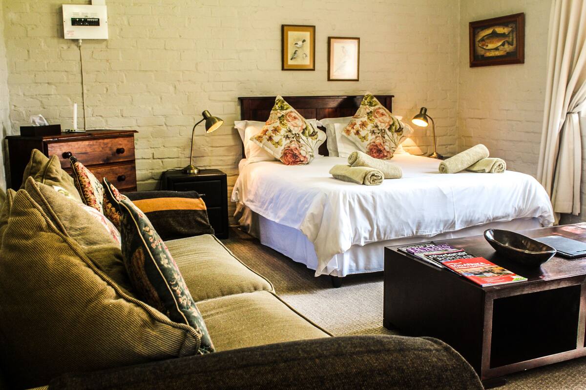 A hotel room with a bed, a sofa, a nightstand, a lamp, and a small table with a bowl and magazines.