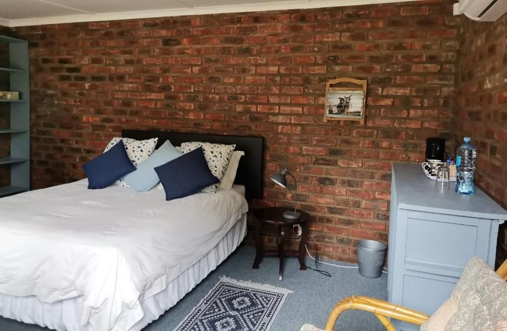 A bedroom with a brick wall, a bed with white bedding, a small side table, a lamp, a water bottle, and a grey dresser.